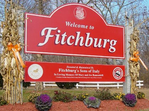 Red Welcome to Fitchburg MA city sign with residential post and rail fencing behind it